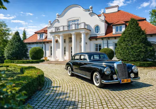 Biały dwór z kolumnowym wejściem i czerwonym dachem, przed którym na brukowanym podjeździe stoi zabytkowy czarny samochód.