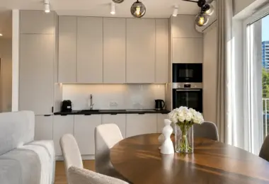 Elegancki apartament Warszawa Wola – jasna kuchnia z zabudową na wymiar, drewnianym stołem jadalnianym oraz dużymi przeszkleniami zapewniającymi naturalne światło.