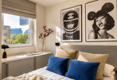 Elegancki apartament Warszawa Wola – nowoczesna sypialnia z tapicerowanym łóżkiem, dekoracyjnymi poduszkami oraz dużym oknem z widokiem na zieleń i zabudowę miasta.
