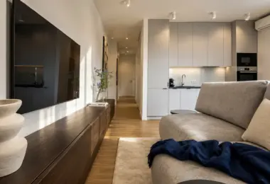 Elegancki apartament Warszawa Wola – nowoczesny salon z designerską sofą, telewizorem, aneksem kuchennym w zabudowie oraz drewnianą podłogą.