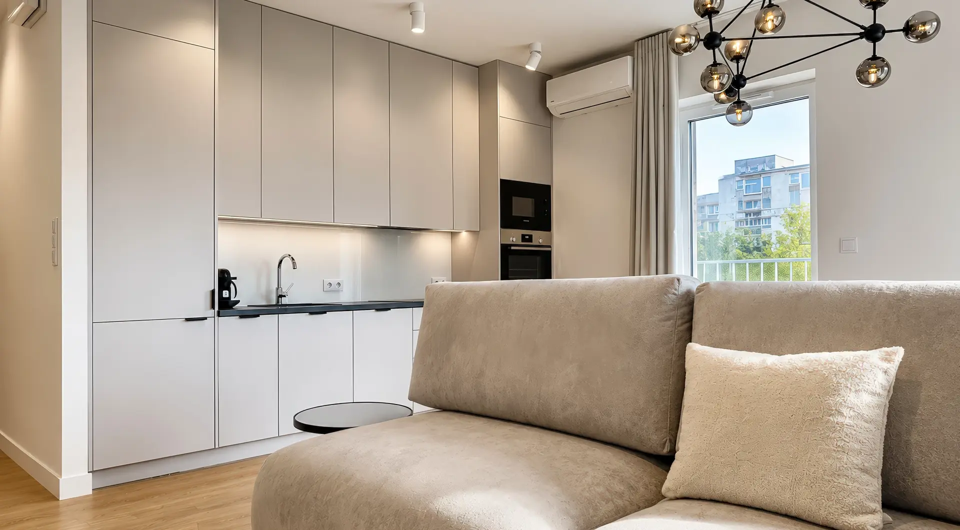 Elegancki apartament Warszawa Wola – nowoczesny salon z designerską sofą, aneksem kuchennym w zabudowie oraz dużym oknem zapewniającym naturalne światło.