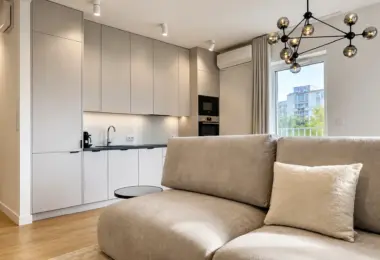 Elegancki apartament Warszawa Wola – nowoczesny salon z designerską sofą, aneksem kuchennym w zabudowie oraz dużym oknem zapewniającym naturalne światło.