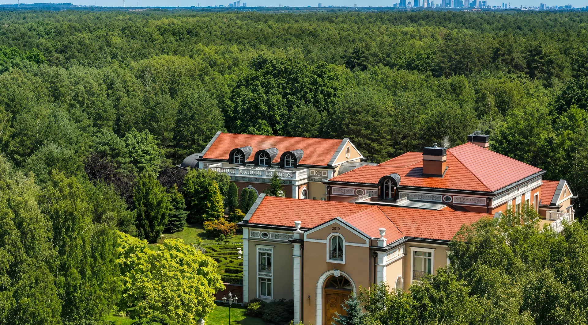 Luksusowa rezydencja pod Warszawą z widokiem z lotu ptaka, prywatnym pałacem otoczonym lasem, czerwonym dachem i panoramą Warszawy w tle